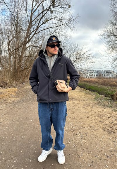 Chaqueta impermeable negra con capucha, sudadera gris debajo, vaqueros holgados azul claro, zapatillas blancas y un pequeño bolso cruzado beige.