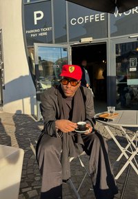 Μαύρο υφασμάτινο μπουφάν και κασκόλ φορεμένα πάνω από ένα μαύρο πουκάμισο. Κόκκινο καπέλο με λογότυπο. Κρατώντας ένα μικρό μαύρο φλιτζάνι καφέ σε στρογγυλό τραπέζι.