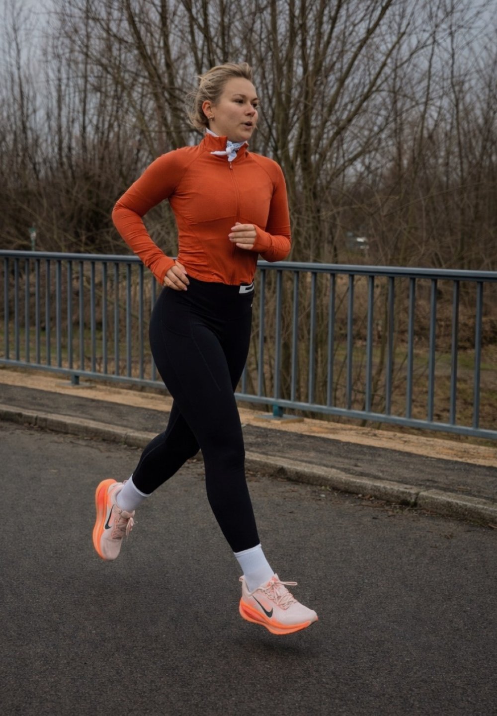 Femme en train de courir, portant un haut ajusté à manches longues orange et un legging noir, avec des chaussures de course Nike roses accentuées de noir et des chaussettes blanches.