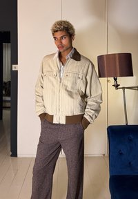 Νέος άνδρας με ξανθά σγουρά μαλλιά, που φοράει μπεζ μπουφάν με φερμουάρ και καφέ διακοσμητικό κορδόνι, καθώς και γκρι παντελόνι, στέκεται σε εσωτερικό χώρο κοντά σε ένα φωτιστικό και μια μπλε καρέκλα.