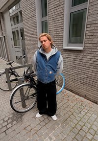 Denimvest over grå hættetrøje, sorte brede bukser og hvide sko. Baggrund: stenbelagt gade med cykler og murstensbygning.