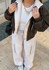 Osoba v bílém žebrovaném tričku, béžových cargo kalhotách, hnědé kožené bundě, vrstvených zlatých řetízcích, prstenech a šedých teniskách stojí na dlaždicové podlaze.