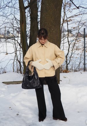 Mujer con chaqueta crema y mitones blancos, sosteniendo un bolso negro, de pie en la nieve junto a un árbol con ramas desnudas.