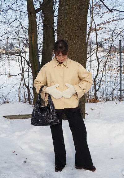 Mujer con chaqueta crema y mitones blancos, sosteniendo un bolso negro, de pie en la nieve junto a un árbol con ramas desnudas.
