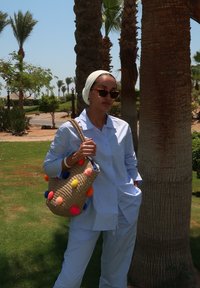 Ανοιχτό μπλε πουκάμισο και παντελόνι από βαμβακερό μείγμα, συνδυασμένα με μια πλεκτή τσάντα με πολύχρωμες φούντες. Φόντο: φοίνικες και πράσινο χορτάρι.