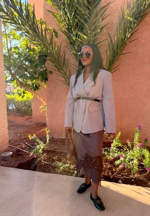 Mujer con blazer beige con cinturón, falda marrón con encaje, zapatos negros, gafas de sol, de pie junto a plantas de palma y flores al aire libre.