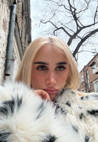 Personne blonde en manteau de fourrure blanc à taches noires reposant le menton sur la main, avec des branches d'arbres nues et des bâtiments en brique en arrière-plan.