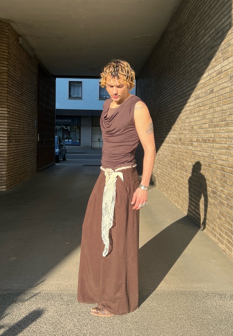 Personne aux cheveux blonds bouclés portant un haut marron sans manches et une longue jupe marron avec une ceinture écharpe blanche, se tenant au soleil et projetant une ombre.