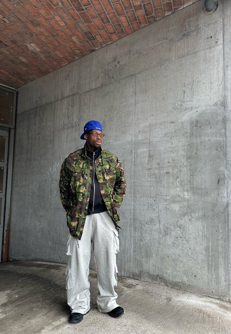 Homme portant une casquette bleue, une veste camouflage, une chemise noire et un pantalon cargo gris, debout contre un mur en béton sous un plafond en briques.