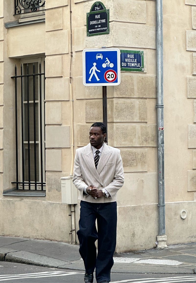 Homme en blazer beige et pantalon foncé marchant au coin d'une rue avec des panneaux de signalisation et des plaques de rue parisiennes visibles.