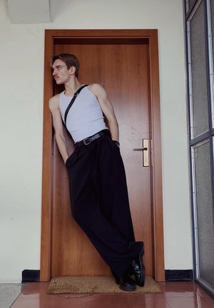 Top blanco sin mangas, pantalones anchos negros y zapatos negros. Se apoya contra una puerta de madera. Texturas suaves, herrajes mínimos y paleta de colores en blanco y negro.