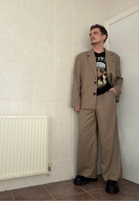 Giacca e pantaloni abbinati a quadri marroni con gambe larghe, indossati sopra una T-shirt grafica. Le calzature sono scarpe nere robuste.