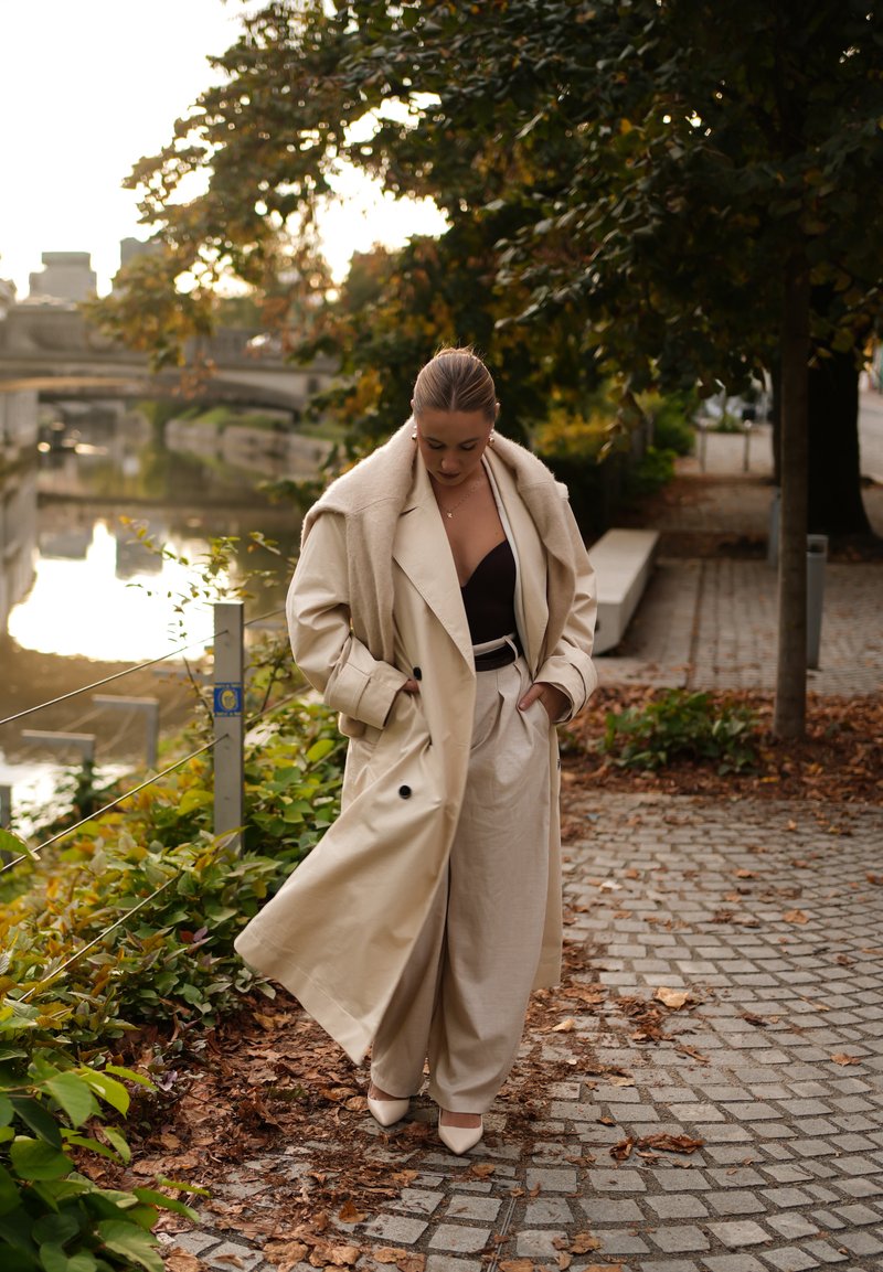 Beige Trenchcoat mit breitem Kragen, getragen über einem dunklen Oberteil und hellen Hosen. Glattes Material; spitze, hochhackige Schuhe runden den Look ab.