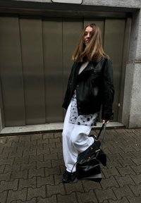 Νεαρή γυναίκα που φοράει μαύρο δερμάτινο μπουφάν και ά σπρο παντελόνι, κρατώντας μια μαύρη τσάντα χειρός, στέκεται σε πλακόστρωτο δρόμο μπροστά σε μεταλλικές πόρτες.