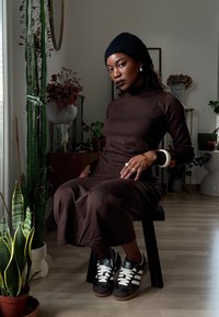 Robe marron à manches longues avec un col haut, associée à des baskets foncées avec des bandes blanches. En arrière-plan, des plantes et des éléments de décoration.
