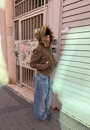 Persona con una chaqueta marrón con capucha forrada de piel y jeans anchos parada en una acera junto a una puerta metálica con graffiti y una persiana cerrada.