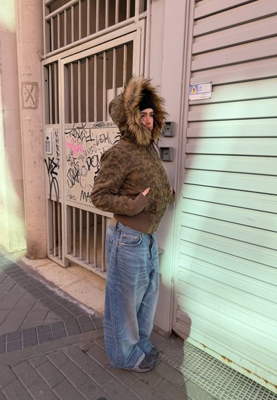 Persona con una chaqueta marrón con capucha forrada de piel y jeans anchos parada en una acera junto a una puerta metálica con graffiti y una persiana cerrada.