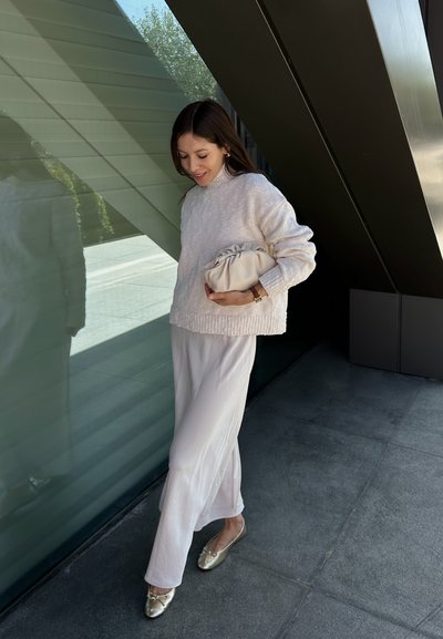 Suéter texturizado en beige claro combinado con pantalones fluidos de crema clara y bailarinas doradas. Sosteniendo un suave bolso de mano color crema.