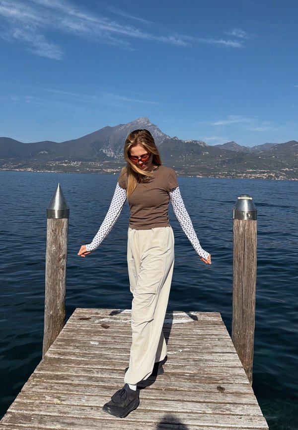 Junge Frau mit Sonnenbrille steht auf einem Holzsteg an einem See mit Bergen im Hintergrund unter klarem blauem Himmel.