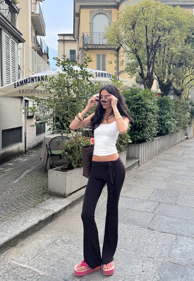 Una mujer está de pie en una calle, vistiendo una camiseta sin mangas blanca, pantalones acampanados negros con un lazo y sandalias de plataforma rosas. Sostiene unas gafas de sol y un bolso de mimbre.