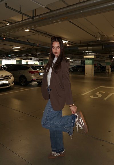 Joven mujer con ropa informal levanta un pie en un parking poco iluminado con coches y pilares de hormigón al fondo.