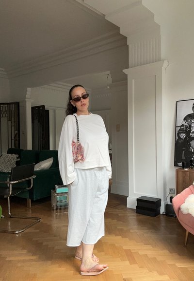 Mujer con camisa blanca de manga larga y pantalones anchos, gafas de sol, sandalias rosas, de pie en un salón con sofá, silla y arte mural.