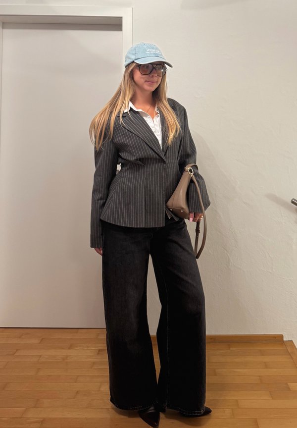 Mujer de pie en el interior, vistiendo un blazer de rayas diplomáticas, camisa blanca, pantalones anchos negros, gorra azul, gafas grandes y sosteniendo un bolso marrón.