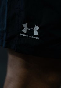 Črne atletske kratke hlače s srebrnim vezenim logotipom Under Armour na robu. Tekstura se zdi gladka in lahka.