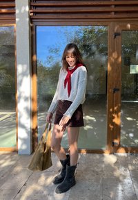 Grijze gebreide trui met een rode stropdas, bruin suedekorte broek, zwarte kniehoge laarzen en een beige tas, staande bij een glazen deur.