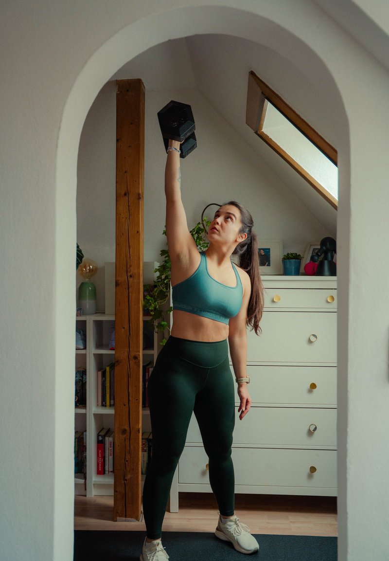 Femme en tenue de sport soulevant un haltère au-dessus de la tête dans une salle de sport à domicile avec un puits de lumière et une poutre en bois en arrière-plan.