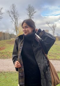 Vrouw draagt een donkergroene waterdichte jas met een geruite voering over een zwarte coltrui, houdt haar kraag vast. Bruine tas over de schouder.