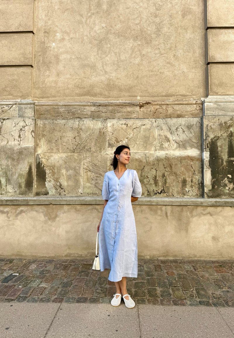 Femme en robe bleu clair boutonnée tenant un sac blanc, debout dans une rue pavée devant un mur en pierre texturé.