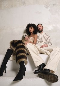 Duas pessoas sentadas num degrau branco. A mulher usa um top fitted bege e um xale de pele listrado. O homem veste uma camisa creme listrada e calças largas creme. Ambos usam calçado escuro.