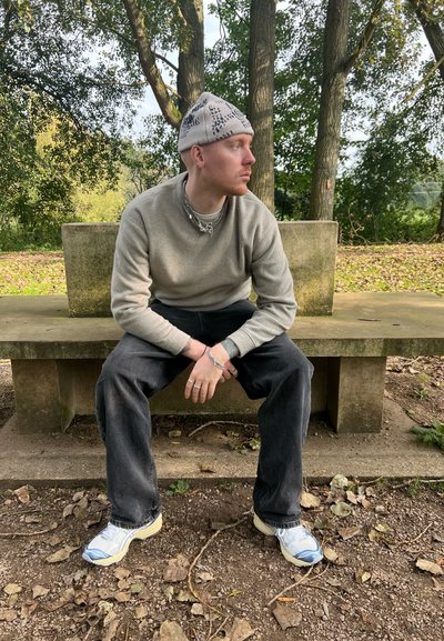 Una persona sentada lleva un sudadera gris claro, jeans oscuros y zapatillas con acentos azules. Tiene un gorro de lana con estampado y collares en capas.