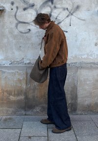 Brun ruskindjakke med elastisk kant, mørkeblå vide jeans og en grå skuldertaske, stående mod en struktureret betonvæg.