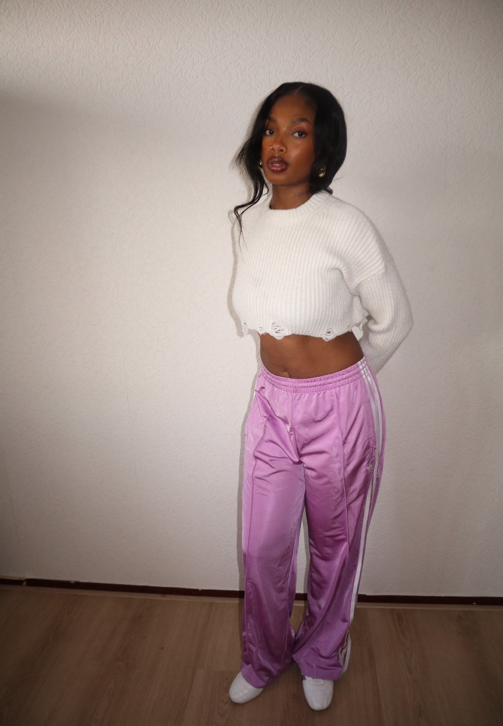 Pull court blanc côtelé avec ourlet effiloché, associé à un pantalon de survêtement violet clair brillant avec des bandes blanches. Le mannequin porte des chaussures blanches.
