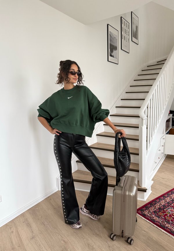 Kvinde iført grøn sweatshirt og sorte læderbukser læner sig mod kuffert med sort håndtaske og står ved trappe i moderne hjem.