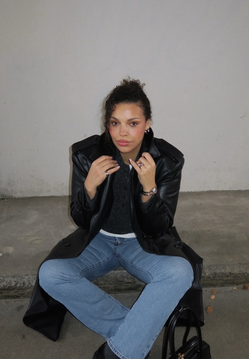 Black leather oversized coat, gray sweater, light blue flared jeans, and jewelry with a textured design, seated on concrete with a black bag.