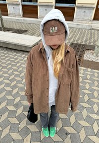 Camisa de pana marrón, sudadera gris con capucha, gorra marrón con logo verde, mochila negra, jeans grises, zapatillas verdes, de pie sobre un pavimento estampado.