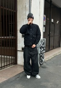 Jeune homme portant une veste matelassée noire, un jean large foncé, des baskets blanches et un bonnet gris orné d'épinglettes, debout sur un trottoir urbain près d'un mur graffiti.
