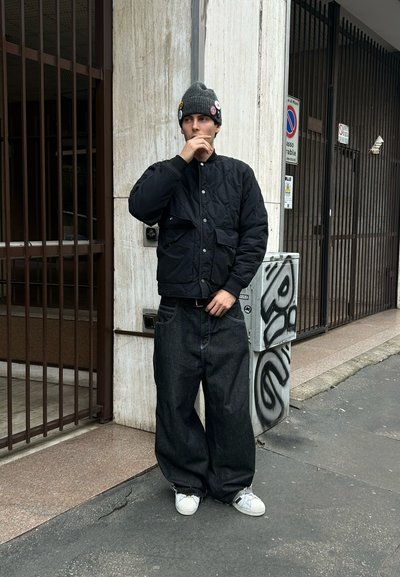 Joven con chaqueta negra acolchada, vaqueros oscuros anchos, zapatillas blancas y gorro gris con chapas, de pie en una acera urbana cerca de una pared con graffiti.