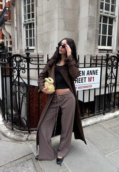 Abrigo marrón, top negro y pantalones de pierna ancha de color marrón claro. Los accesorios incluyen gafas de sol negras y un bolso escultural de color amarillo claro.