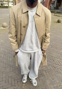 Beige trenchcoat över en grå sweatshirt och matchande sweatpants. Grå sneakers med vita accenter. Guldig kedja synlig runt halsen.