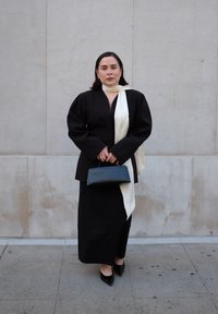 Blazer nero sartoriale con maniche a sbuffo, abbinato a una lunga gonna nera. Una sciarpa bianca pende attorno al collo, sostenendo una borsa nera.