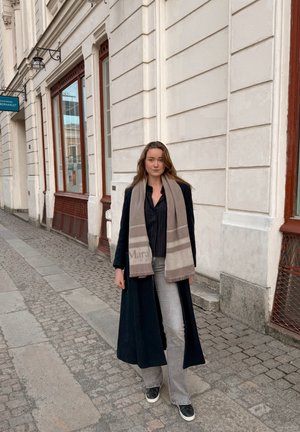 Mujer joven con un abrigo largo negro, bufanda beige, vaqueros grises y zapatillas negras, de pie en una calle adoquinada cerca de un edificio blanco.