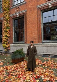 Brun dobbelspent kåpe båret over en lys topp, stående på en bakke dekket av fargerike høstblader. Rød murvegg og vinduer i bakgrunnen.