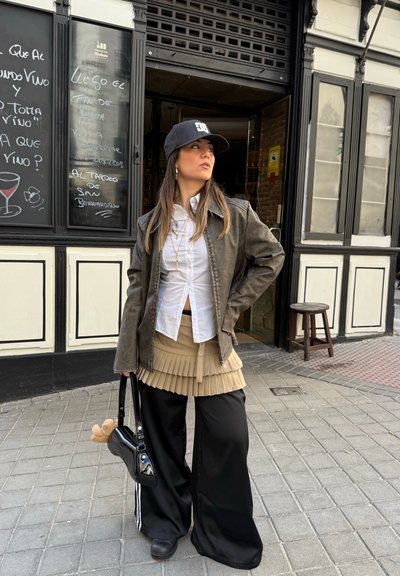 Una mujer con una chaqueta de cuero marrón sobre una camisa blanca, una falda plisada beige y pantalones anchos negros, sosteniendo un bolso negro.