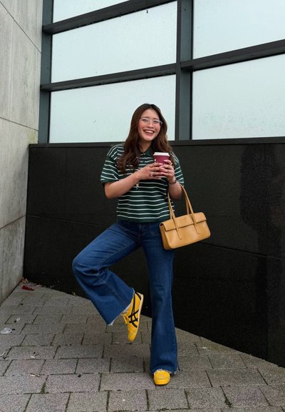 Joven mujer sonriendo, sosteniendo una taza de café, con camisa de rayas, vaqueros azules, zapatillas amarillas y llevando un bolso marrón claro junto a la pared de un edificio.