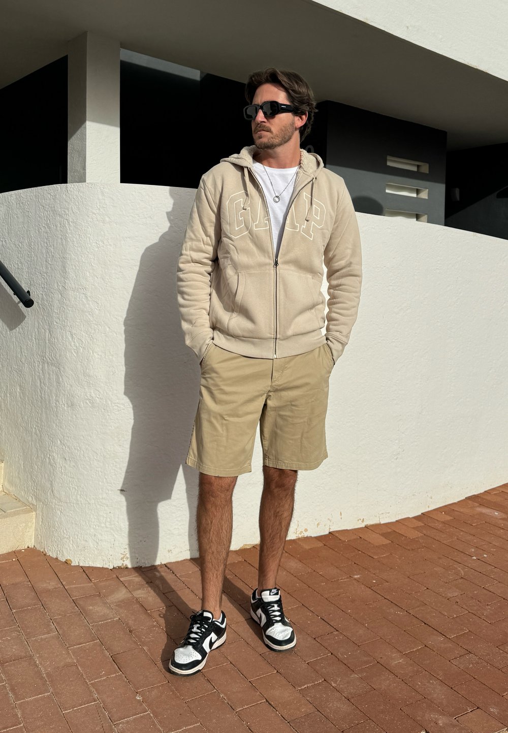 Beige zip-up hoodie with GAP logo, white t-shirt, khaki shorts, and black sneakers with white accents, standing on brick pavement against white wall.