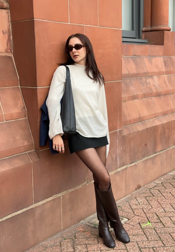 Femme en lunettes de soleil appuyée contre un mur de briques rouges, portant un pull blanc, une jupe noire, des collants transparents, des bottes marron, et portant un sac à bandoulière noir.